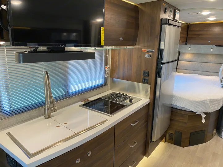 Inside a modern RV rental: complete kitchen with stove, sink, and fridge, plus TV and comfy bed area.. Winnebago View 2022