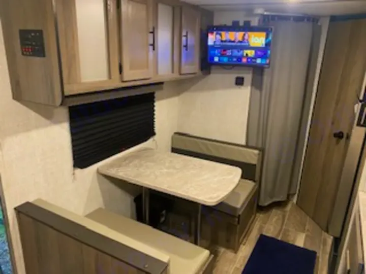 RV rental dinette with cozy bench seating, table, and wall-mounted TV. Overhead cabinets and a window enhance this compact camper space.. Gulf Stream Enlighten 2022