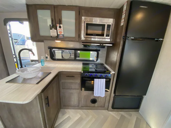 Modern rv rental kitchen with L-shaped counter, sink, microwave, stove, oven, and full-size black refrigerator.. Forest River Wildwood X-Lite 2022