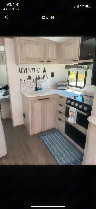 Modern RV rental kitchen: light wood cabinets, white countertops, black sink/stove/microwave, subway tile backsplash, and 'Adventure Awaits' decal.. Shadow Trailers 277bhs 2022