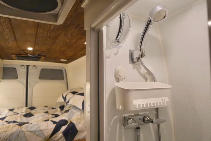Compact shower with handheld head and mirror, beside a cozy bed with patterned bedding in a modern rv rental.. Mercedes-Benz Sprinter RV Motorhome Campervan 2022