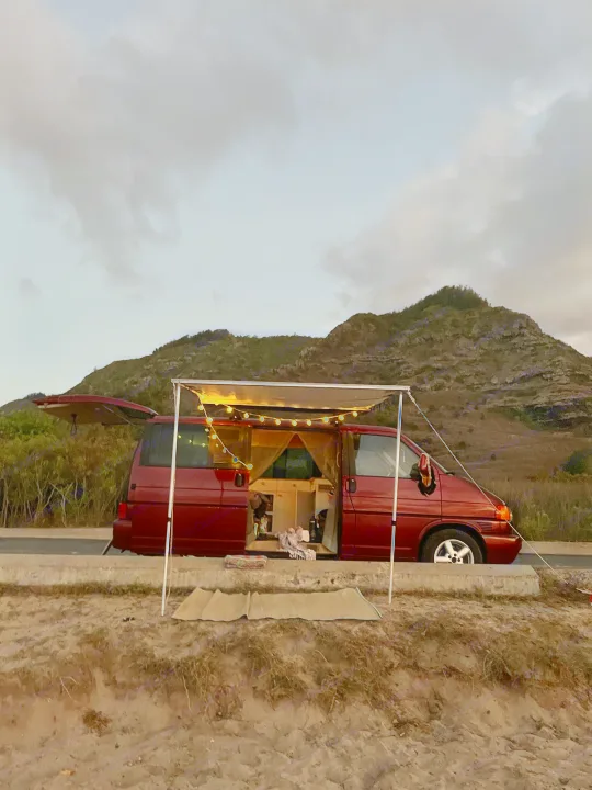 Red camper van RV rental with illuminated awning set for cozy evening camping by sandy beach, mountains in distance.. Volkswagen Eurovan 2003