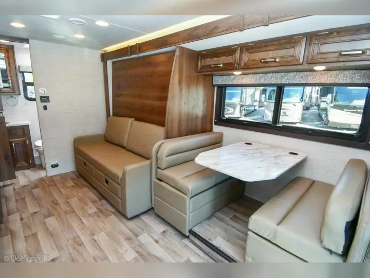 Inside an RV rental: living area with light wood floor, convertible sofa, and dinette. Large windows show other RVs outside.. Jayco 24M 2022