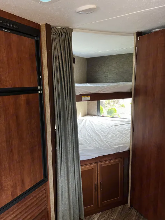 RV rental interior showing bunk beds with a gray privacy curtain, a Dometic refrigerator, and wood cabinets.. Keystone RV Passport Grand Touring 2016