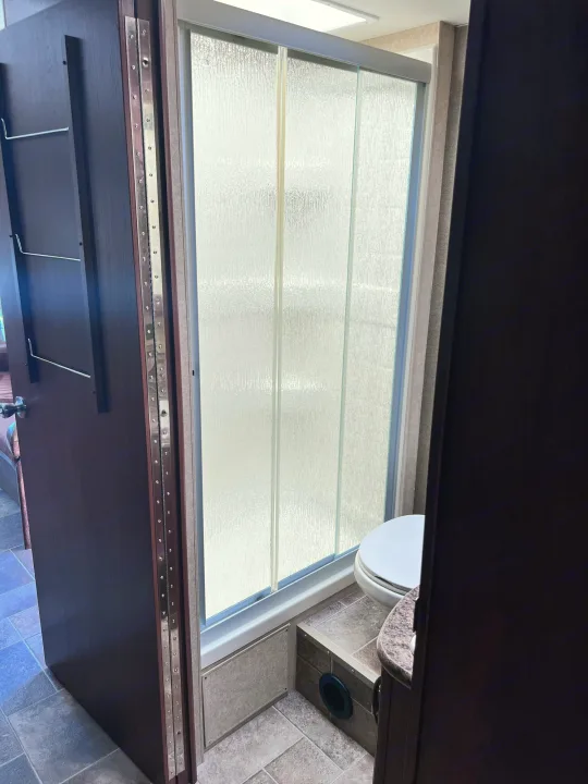 Compact RV rental bathroom features a glass shower, toilet, and a hinged door with hanging storage, optimizing space for travel.. Thor Freedom Elite 2015