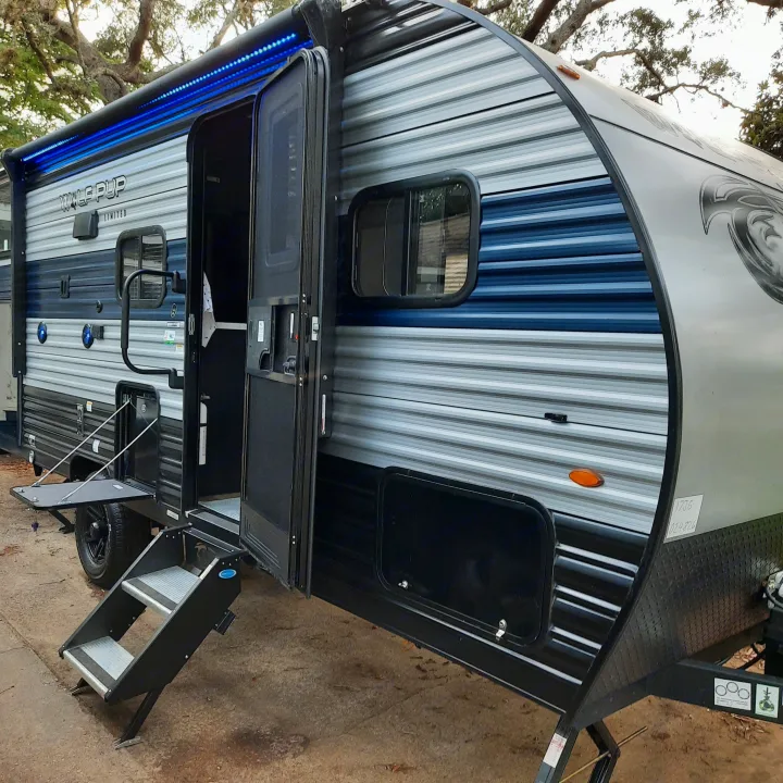 Modern gray and blue rv rental with bright blue LED awning lights, open door, and steps. Parked in a natural setting.. Forest River Cherokee Wolf Pup 2022
