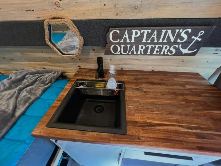 Inside this rv rental: a black sink on a wooden counter, with a bed nearby. Nautical 'Captain's Quarters' sign and rope mirror. Mobile living comfort.. Ford Econoline E350 Extended 2004