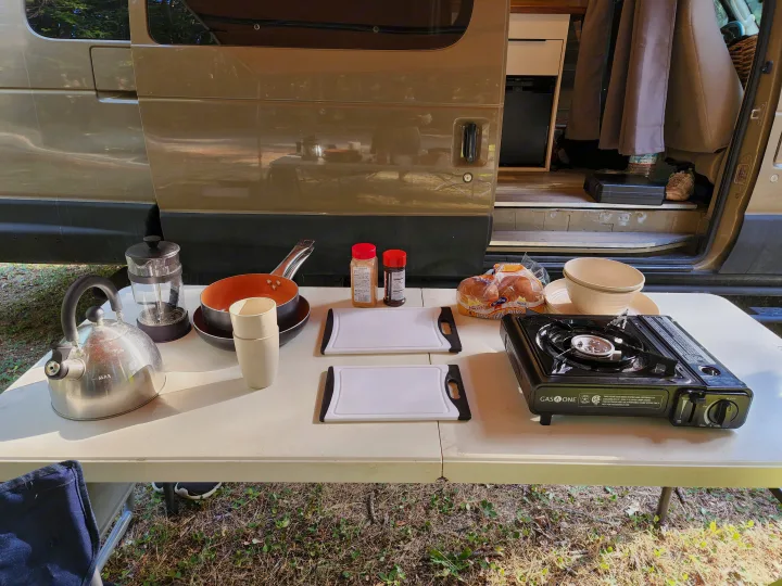 An outdoor cooking setup for an RV rental, featuring a camp stove, kettle, and food items next to a camper van.. Ford Econoline E350 Extended 2004