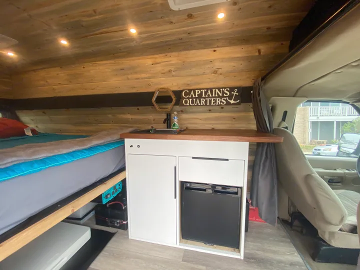 RV rental interior: cozy bed, wood-paneled walls, and kitchenette with sink and fridge. A "Captain's Quarters" sign adorns this camper van.. Ford Econoline E350 Extended 2004
