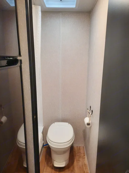 RV rental bathroom featuring a white toilet, wood accent, unique rope TP holder, and glimpse of shower.. Forest River Cherokee Wolf Pup 2022