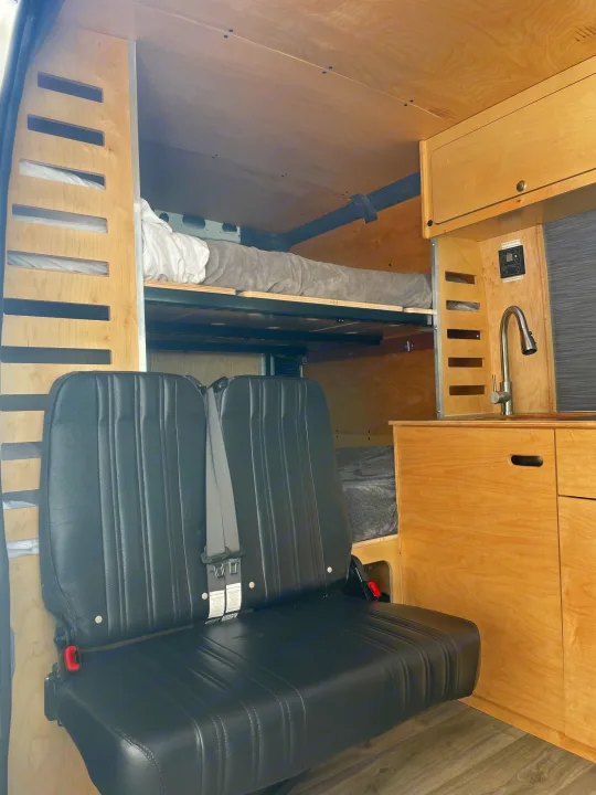 Inside this modern rv rental: bunk beds, a kitchen with sink, and a multi-passenger bench seat in a wood-paneled camper van.. RAM ProMaster 2022