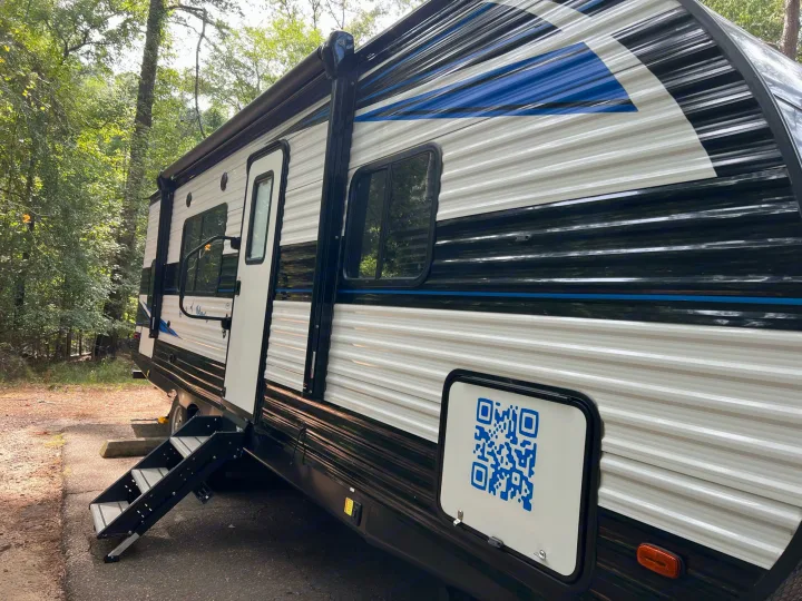 An RV rental, with its entry steps down and a QR code on its side, is parked in a lush, green campsite.. Heartland Prowler 2021