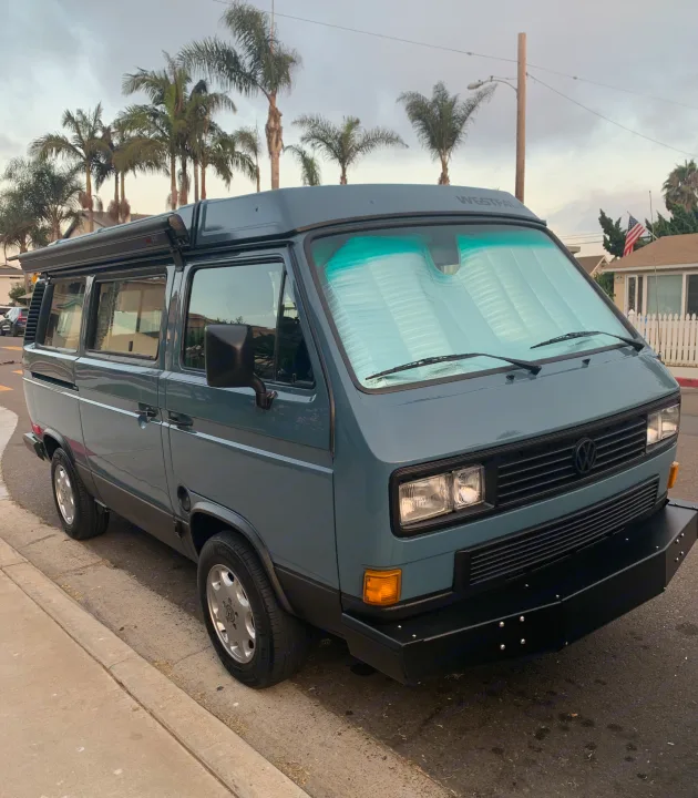 A blue-grey vintage Volkswagen Westfalia camper van, ideal for an RV rental, is parked on a palm-lined street.. Volkswagen Westfalia 1990