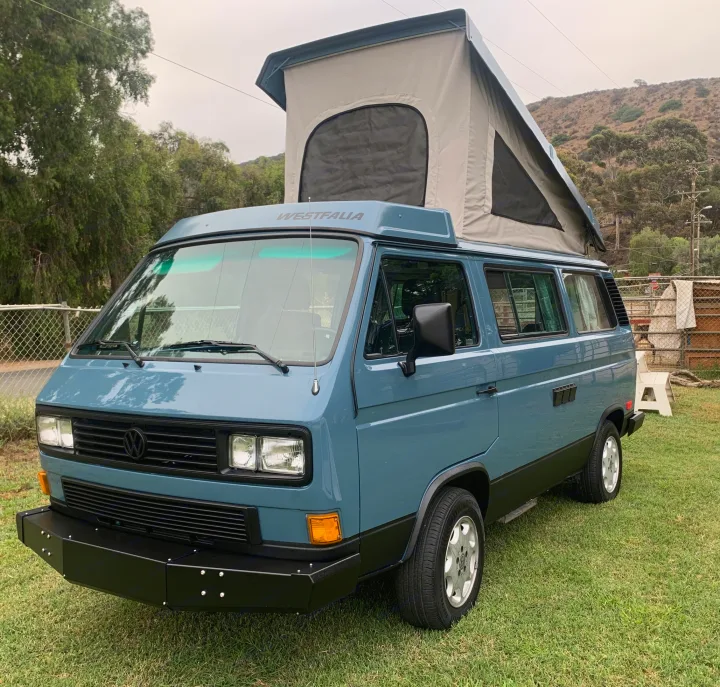 A striking blue VW Westfalia pop-top camper van, perfect for an rv rental, parked on green grass with hills in the background.. Volkswagen Westfalia 1990