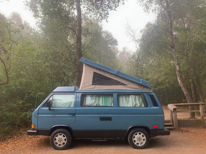 Blue VW camper van with its pop-top extended, parked on a misty, tree-lined road. Explore with an RV rental!. Volkswagen Westfalia 1990