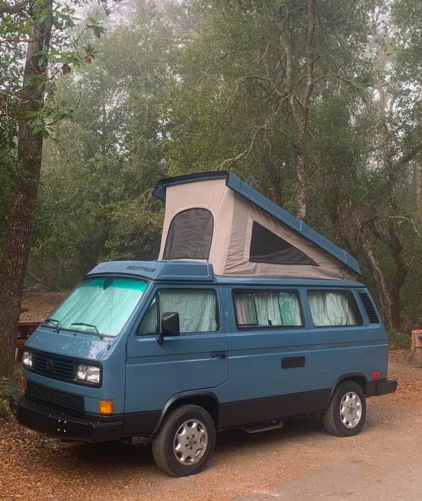 Blue VW Westfalia pop-top camper van parked in misty woods. Ideal for your next rv rental adventure and camping trip.. Volkswagen Westfalia 1990