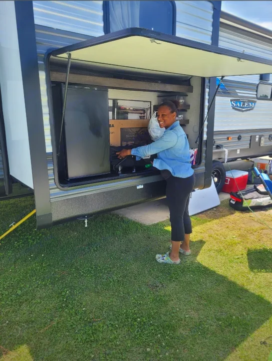A smiling woman sets up her RV rental's outdoor kitchen, complete with a fridge and grill, for camping fun.. Forest River Salem 2020