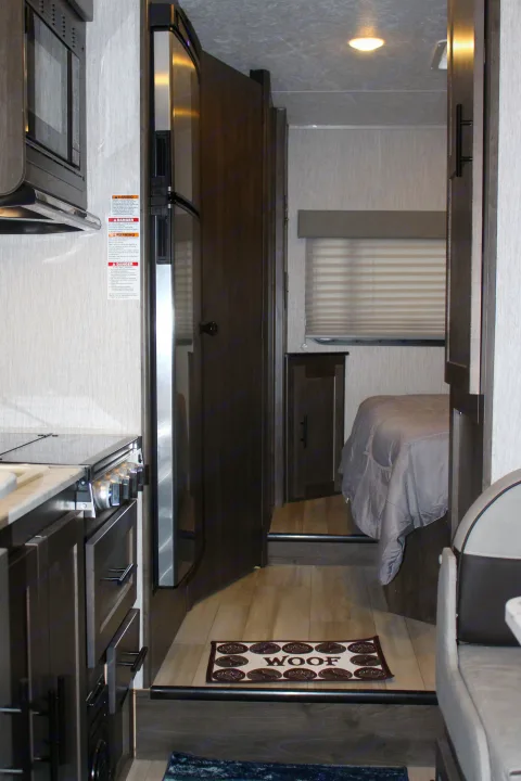 This RV rental interior showcases a compact kitchen, refrigerator, and a hallway leading to the bedroom, with a "WOOF" mat.. Forest River Coachmen Leprechaun 2022
