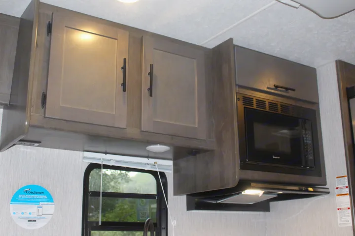 An RV rental interior shows a kitchen with dark wood-look upper cabinets, an integrated microwave, and a window.. Forest River Coachmen Leprechaun 2022