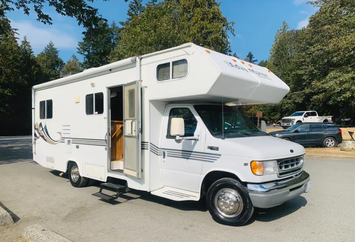 A white Adventurer Class C motorhome RV rental is parked on asphalt with trees in the background under a blue sky.. Slumber Queen Adventurer 2001