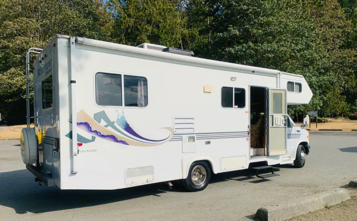 White Class C RV rental parked outdoors. Features include an open door, a ladder, and a rear spare tire. Ready for adventure!. Slumber Queen Adventurer 2001