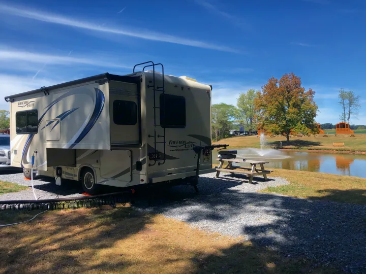 A spacious RV rental is parked on a gravel site by a scenic pond with a fountain and a wooden picnic area.. Thor Freedom Elite 2018