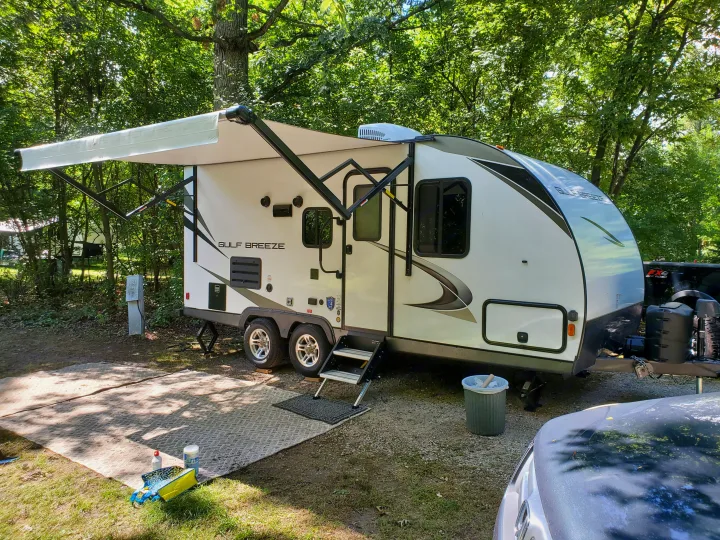 This RV rental, a Gulf Breeze travel trailer, is parked at a wooded campsite with its awning extended and hookups connected.. Gulf Stream 21QBS 2022