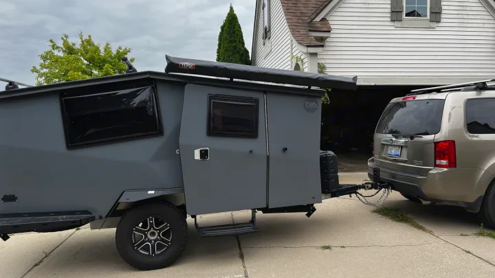 A rugged gray camper trailer, a perfect RV rental option, is hitched to a gold SUV in a driveway, ready for outdoor adventures.. TAXA Cricket Overland Edition 2020