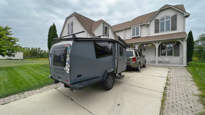 A gray Cricket camper, perfect for an RV rental, sits in a driveway next to an SUV at a large house.. TAXA Cricket Overland Edition 2020