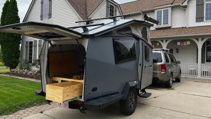 Gray teardrop camper with open rear kitchen/storage, hitched to an SUV in a driveway. Perfect RV rental for adventures!. TAXA Cricket Overland Edition 2020