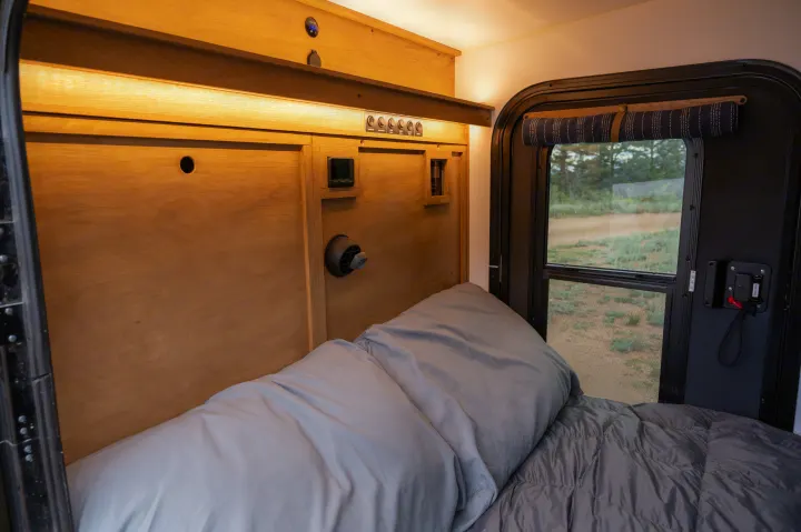 This RV rental offers a cozy bed with light grey bedding, warm wooden walls, essential controls, and a window view.. White River X Camper Trail Explorer 2021