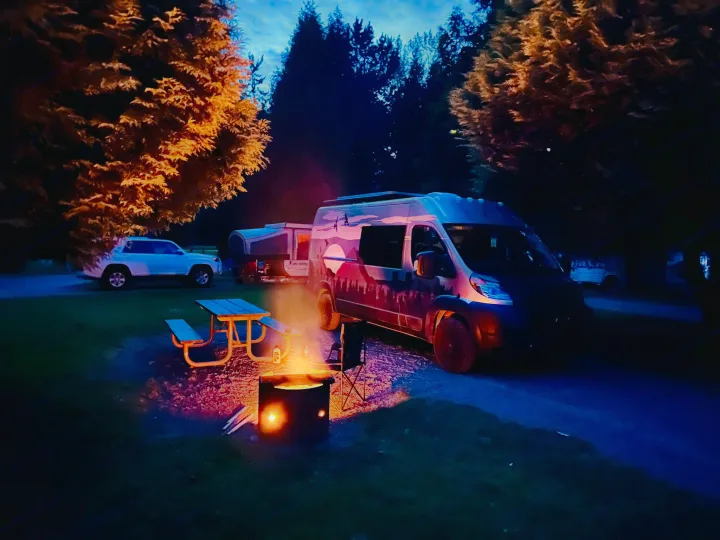 Cozy night at a campsite with an RV rental, glowing campfire, picnic table, and camper van under large trees.. Dodge Ram 1500 2020