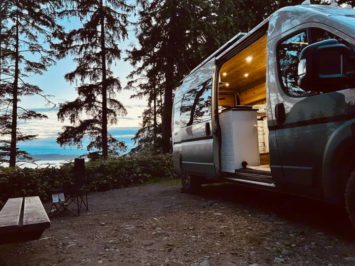 An RV rental, door open to a lit interior, overlooks the calm ocean at dusk. Parked among tall trees, a bench and chair complete this scenic camp.. Dodge Ram 1500 2020
