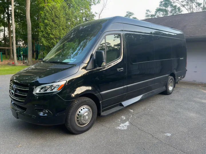 A shiny black Mercedes-Benz Sprinter van, often used for RV rental, parked on an asphalt driveway by trees & a house.. Ultimate Toys Ultimate Coach 2022