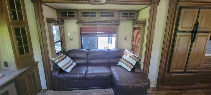 Spacious RV rental living area with a brown leather sectional, windows, and wood cabinets.. Keystone RV Alpine 2015