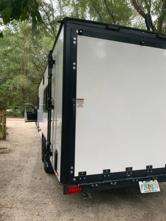 White RV rental trailer parked on a dirt path in a wooded campsite, displaying its back door, Florida plate, and side ladder.. Forest River no boundaries 2020