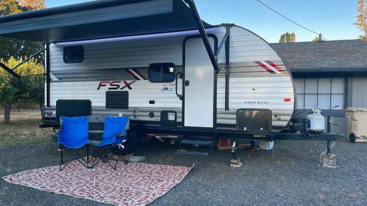A spacious RV rental featuring an extended awning with LED lights, blue chairs, and a patterned mat for outdoor relaxation.. Forest River Wildwood FSX 2021