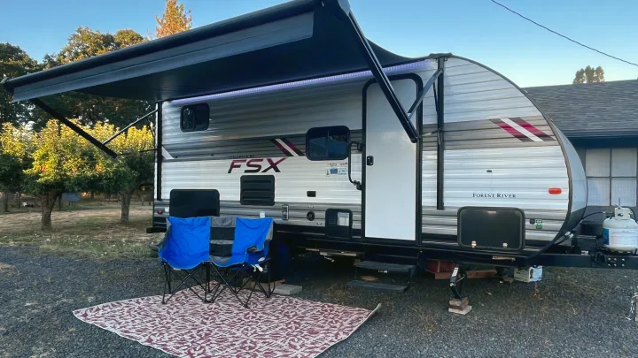 Enjoying an RV rental setup: a travel trailer with an extended awning, glowing lights, chairs, and a rug on gravel.. Forest River Wildwood FSX 2021
