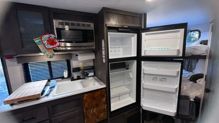 RV rental interior featuring a modern kitchen: open Dometic refrigerator, microwave, double sink, and bunk beds.. Forest River Wildwood FSX 2021