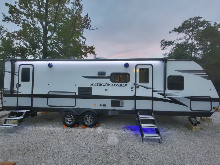 Brightly lit Jay Feather travel trailer at dusk on a gravel RV rental site, showing exterior white & blue LED lights.. Jayco Jay Feather 2023