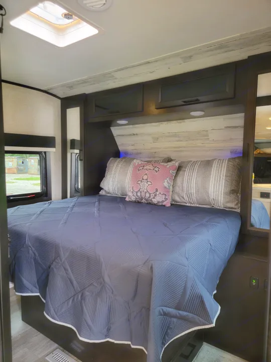 Inside an RV rental: a neat bedroom featuring a blue quilted bedspread, dark cabinetry, and a bright overhead skylight.. Jayco Jay Feather 2023