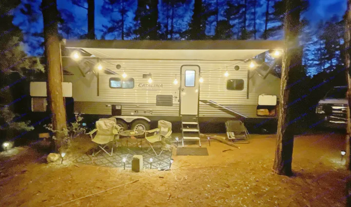 Cozy rv rental campsite at night, bathed in the glow of string lights. Features a modern travel trailer, chairs, and ground lighting.. Forest River Coachmen Catalina Legacy 2022