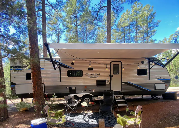 This RV rental campsite features a modern travel trailer with an awning, string lights, and comfy outdoor chairs under tall trees.. Forest River Coachmen Catalina Legacy 2022
