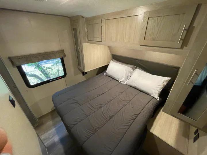 Cozy RV rental bedroom featuring a bed with dark grey bedding and white pillows, light wood cabinets, and a window with tree views.. Forest River Coachmen Catalina Legacy 2022