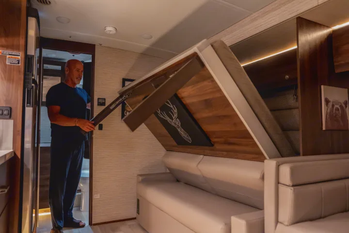 A man converts an RV rental living area by pulling down a clever, space-saving wall bed above a beige sofa.. Winnebago View 2025