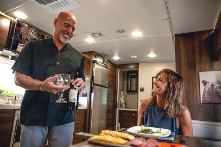 Happy couple sharing wine and charcuterie in their modern RV rental. Spacious interior with kitchen & TV. Perfect for travel!. Winnebago View 2025