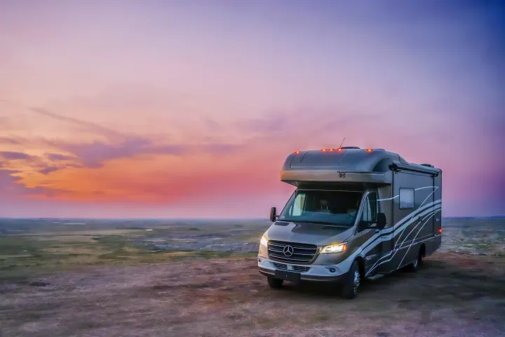 A modern two-tone rv rental is parked at sunset, with headlights on, under a vibrant pink-purple sky in a vast landscape.. Winnebago View 2025