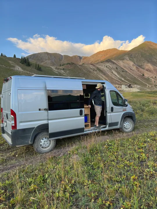 Scenic RV rental adventure: a man stands in a silver camper van, parked on a dirt path in grassy mountains.. Dodge Promaster 2500 2019