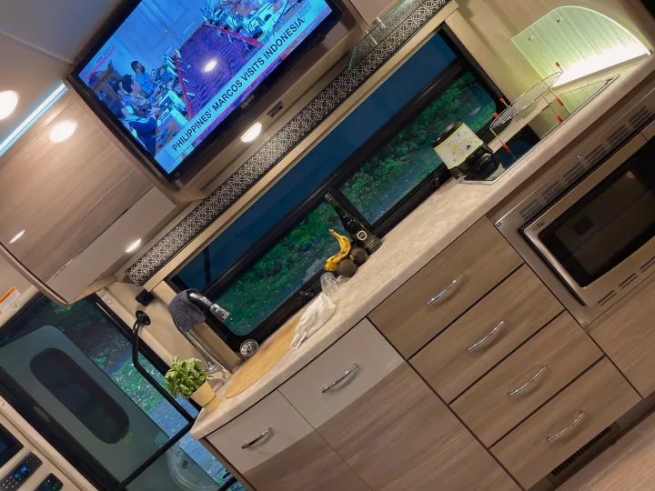 An RV rental's compact kitchen and living area, complete with a TV showing news, sink, microwave, and ample storage for travel.. Thor Tiburon 2020