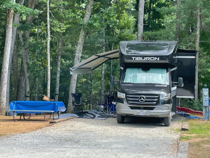 Black Tiburon RV rental parked at wooded campsite 725 with awning extended, chairs, and a blue-tarped picnic table.. Thor Tiburon 2020